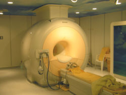 Room-high yellow-grey medical machine with a man-size hole in the middle and a stretcher directly in front of it