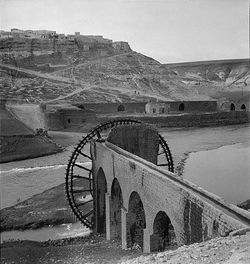 الرستن (على ربوة في الخلفية) وناعورة/ساقية (في المقدمة) يفصلهما نهر العاصي، عقد 1930