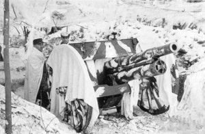 A cannon wears a white blanket on the snow and two men pose side of it.