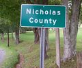Several markers designating the border between Nicholas and Greenbrier counties in West Virginia, USA along a secondary road. Notice the older stone survey markers a few meters behind the modern highway sign.