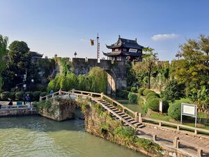 Stone footbridge spanning a canal in front of an ancient city wall and a traditional Chinese gate tower under a clear sky.